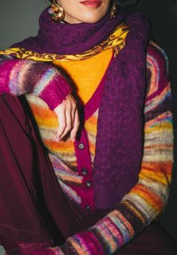 Donna con grandi orecchini dorati, una sciarpa viola lavorata a maglia, un maglione a righe colorate e soffice, pantaloni bordeaux e una maglietta gialla, seduta contro uno sfondo scuro.