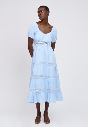 Frau steht mit den Armen hinter dem Rücken, trägt ein hellblaues Midikleid mit Puffärmeln und braune Sandalen, leicht nach rechts gewandt.