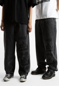 Jeans in denim grigio scuro, larghi, con stampa grafica nera, abbinati a scarpe neri e bianche. Le magliette sono rispettivamente nere e bianche.