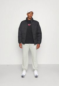 Sort dunjakke med quiltet design, brukt over en svart t-skjorte med rødt logo. Lysegrå joggebukser og hvite joggesko fullfører antrekket.