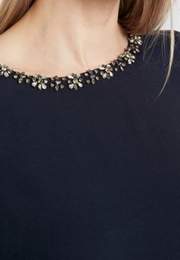 Nahaufnahme des Ausschnitts einer Frau, der mit filigranen floralen Verzierungen aus Silber, Gold und klaren Steinen auf einem dunkelblauen Stoff geschmückt ist.