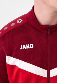 Rode sportieve jas met hoge kraag, ritssluiting, witte streep, reliëfpatroon en "JAKO" logo op de borst. Glad textuur van de stof.
