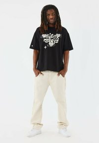 Zwarte t-shirt met witte grafische tekst "PROJECX PARIS," gecombineerd met beige broek met rafelige naden, afgemaakt met witte sneakers.