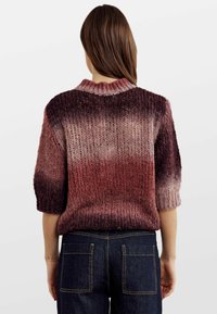 Žena v volném pleteném svetru s krátkým rukávem v gradientových odstínech burgundy, doplněném o tmavě modré džíny s velkými zadními kapsami.