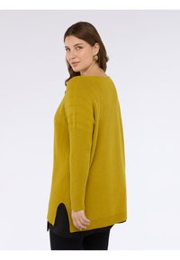 Maglione oversize giallo senape con texture a coste, caratterizzato da scollo tondo, spacchi laterali e vestibilità comoda, abbinato a un top nero sotto.