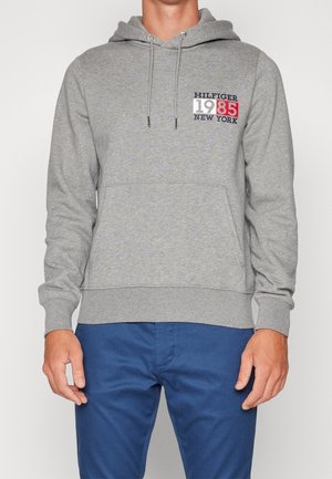 Sweat-shirt à capuche gris avec une poche kangourou, arborant un imprimé graphique rouge et bleu comprenant "HILFIGER 1985 NEW YORK".