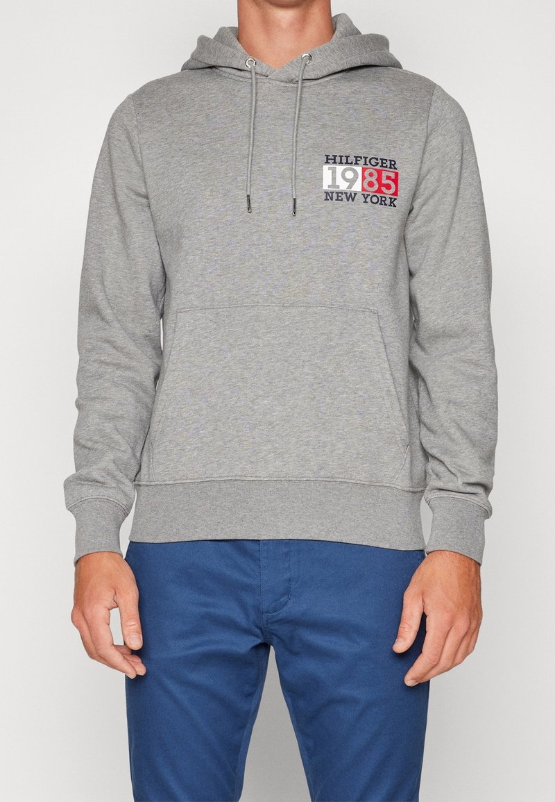 Felpa grigia con cappuccio e tasca a marsupio, caratterizzata da una stampa grafica rossa e blu che include "HILFIGER 1985 NEW YORK."
