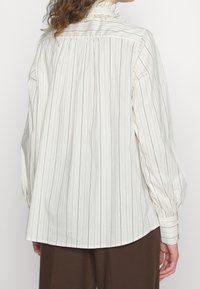 Vit långärmad skjorta med tunna vertikala ränder, volangkrage och elastiska ärmar; lättviktsmaterial, avslappnad passform, rundad nederkant.