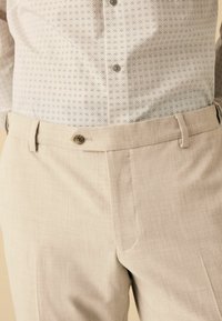 Beige Hosen mit einer glatten Textur, die einen Knopfverschluss und Gürtelschlaufen aufweisen. Der Stoff hat ein dezentes Webmuster.