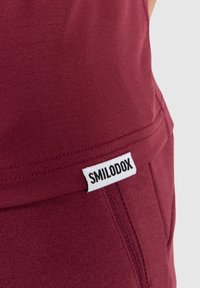 Burgundy träningsshorts tillverkade av stretchigt material. Har en synlig vit "SMILODOX" logotyp och sömmar längs kanterna.