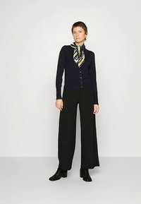 Marineblauwe vest met knopen, gecombineerd met wijde zwarte broek. Geaccentueerd met een sjaal met een patroon in groen- en blauwtinten.