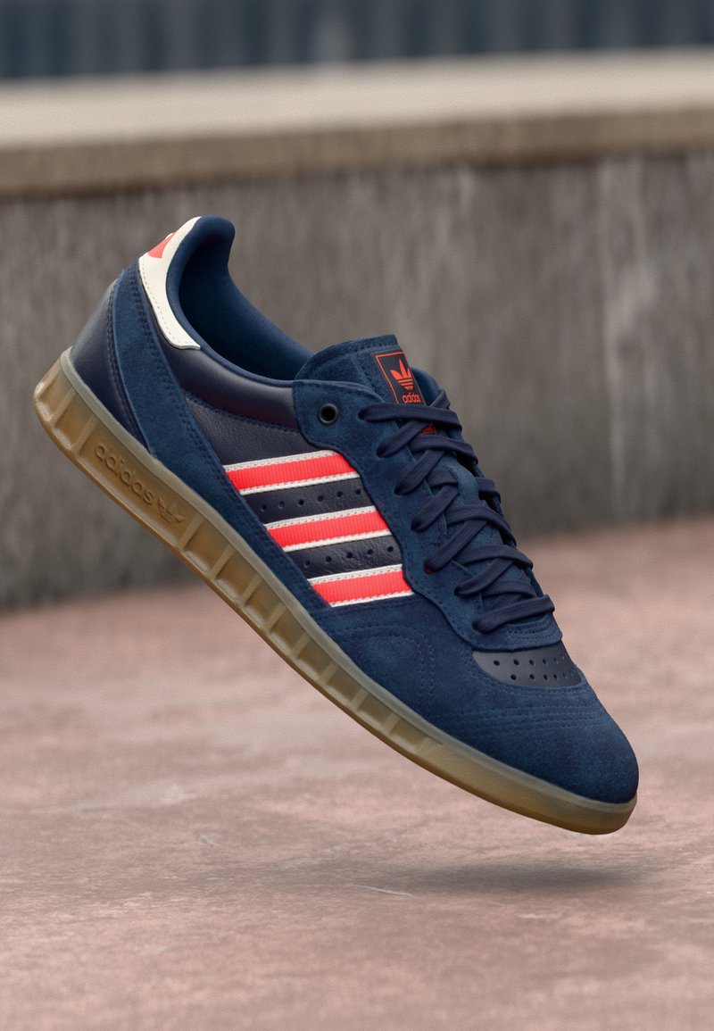 Mörkblå Adidas-sneaker med röda och vita ränder, gummisula och snören, sväva ovanför en rosa betongyta.