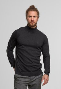 Mann mit Bart und zu einem Zopf gebundenem Haar trägt einen dunkelgrauen Rollkragenpullover und graue gemusterte Hosen, steht vor einem schlichten hellen Hintergrund.