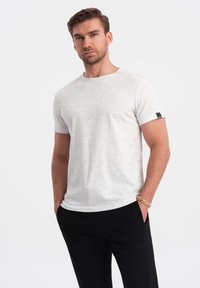 Hvid t-shirt med sorte spidser, rund halsudskæring, korte ærmer og et lille sort mærke på venstre ærme, parret med sorte bukser.