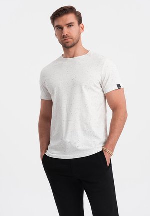 White t-shirt with black speckles, round neckline, short sleeves, and a small black label on the left sleeve, paired with black pants.