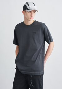 Nagyméretű sötétszürke póló kerek nyakkivágással és kis logóval. Fekete farmerral párosítva, szegecses díszítéssel.