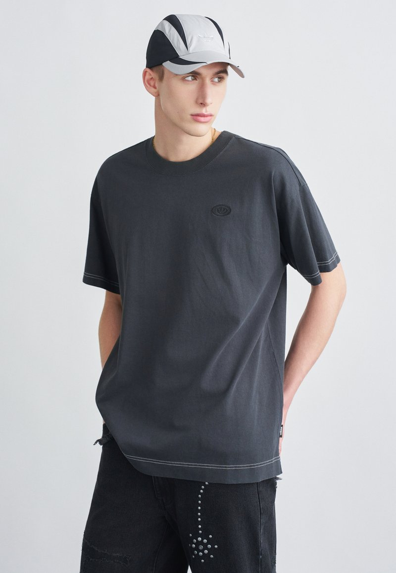 Nagyméretű sötétszürke póló kerek nyakkivágással és kis logóval. Fekete farmerral párosítva, szegecses díszítéssel.