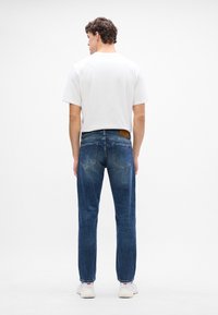 Jeans in denim blu con taglio dritto, dotati di tasche posteriori e una patch in pelle marrone nella waistband, indossati con una t-shirt bianca semplice.
