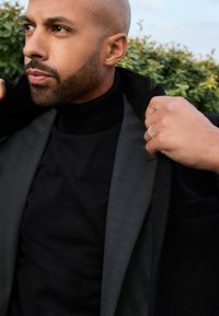 Jersey de cuello alto negro bajo un blazer gris con un patrón sutil, solapas texturizadas y una mano ajustando el cuello. Follaje verde en el fondo.