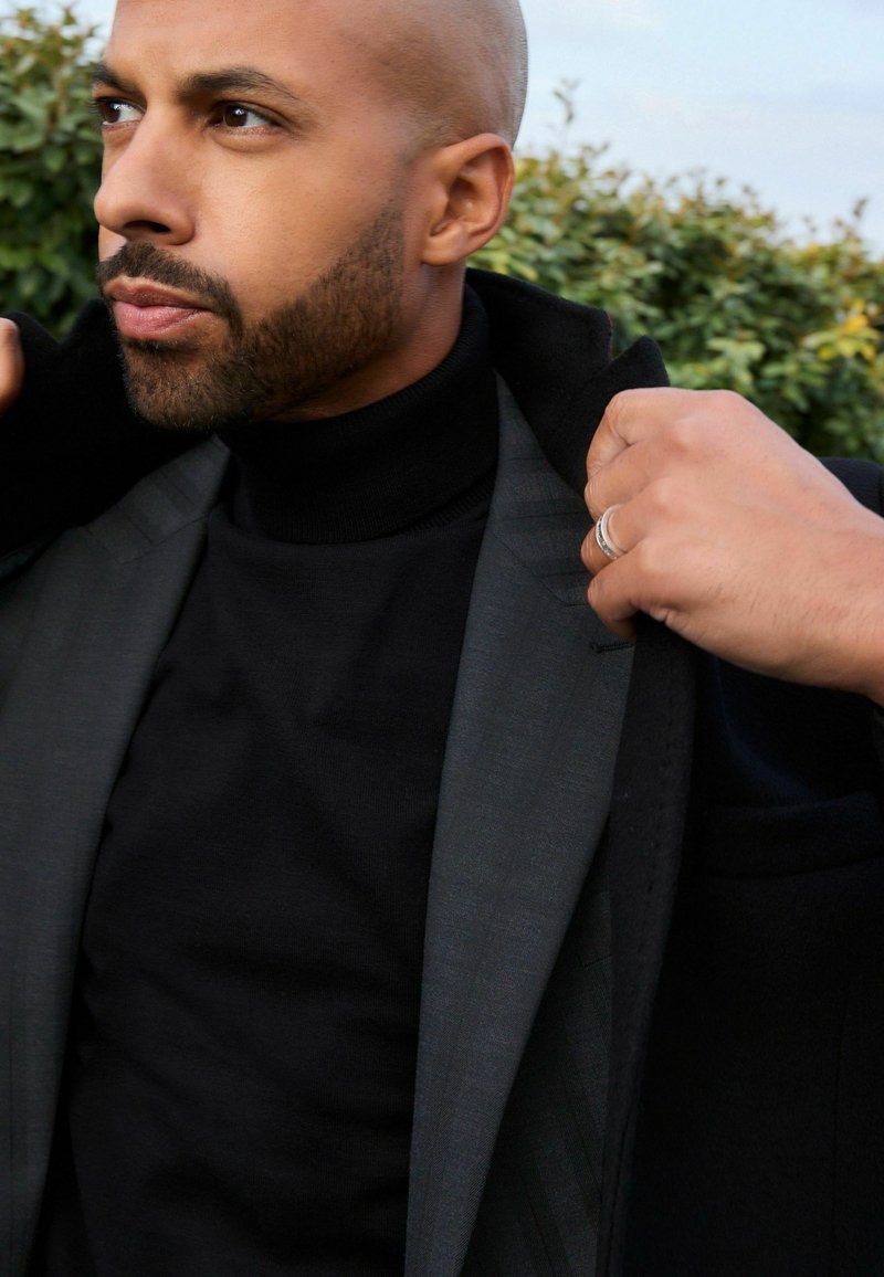 Jersey de cuello alto negro bajo un blazer gris con un patrón sutil, solapas texturizadas y una mano ajustando el cuello. Follaje verde en el fondo.