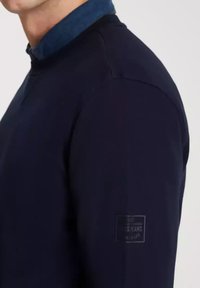 Granatowy sweatshirt z teksturowanej tkaniny, z prążkowanym kołnierzem i subtelną naszywką z logo na rękawie. Posiada dopasowany krój i casualowy fason.