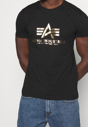 Mann trägt schwarzes T-Shirt mit goldenem Alpha Industries-Logo und Schriftzug, kombiniert mit blauen Jeans, steht vor weißem Hintergrund.