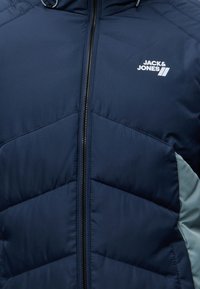 Veste matelassée bleu marine avec fermeture éclair noire et logo blanc "Jack & Jones" sur la poitrine gauche, avec un panneau latéral gris clair.