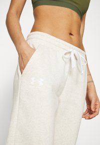 Lichtgrijze trainingbroek met een trekkoord in de taille, zijzakken en een wit Under Armour-logo op het linkerpoot. Zachte stoftextuur.