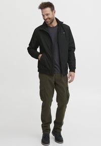 Črna lahka jakna z visokim ovratnikom, oblečena čez temno sivo majico. Oljčno zelene cargo hlače s stranskimi žepi in črne čevlje.