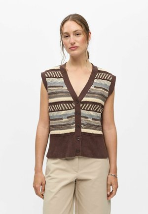 Femme portant un gilet sans manches en maille marron, beige et gris à motifs, boutonné, et un pantalon beige, debout devant un fond blanc uni.
