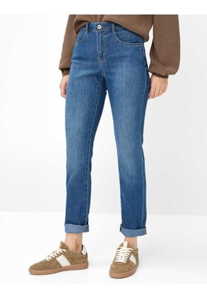 Person mit hochgekrempelten blauen Jeans, beigen und weißen Sneakers und braunem Langarmpullover, stehend auf einem weißen Boden.