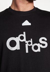 Svart bomullströja med en stor vit Adidas-logga, med en mindre vit logga ovanför. Slät textur, rundad halsdesign.