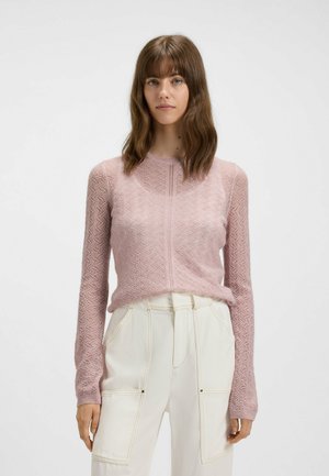Maglione lavorato a maglia rosa chiaro con un motivo a zigzag testurizzato, scollo rotondo e maniche lunghe, abbinato a pantaloni a vita alta di colore bianco sporco.