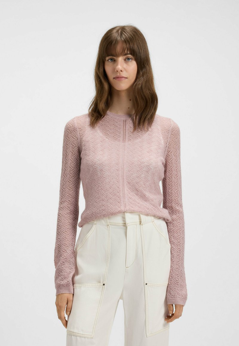 Hellrosa gestrickter Pullover mit einem strukturierten Zickzack-Muster, rundem Ausschnitt und langen Ärmeln, kombiniert mit off-white hochgeschnittenen Hosen.