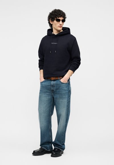 Homme portant un sweat à capuche noir avec un petit logo, un jean bleu, une ceinture marron, des chaussures noires et des lunettes de soleil foncées, debout les mains dans les poches.