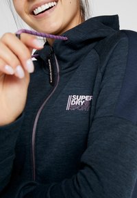 Sudadera con cremallera en color azul marino, hecha de tejido texturizado, con una cremallera y un logotipo en color púrpura. La capucha cuenta con un cordón ajustable con acentos púrpuras.