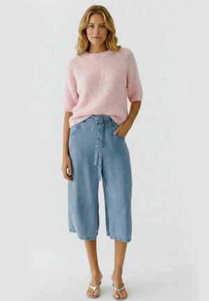 Vrouw draagt een lichtroze zachte trui met halve mouwen, lichtblauwe high-waisted denim culottes en zilveren sandalen met open tenen, staat met één hand in haar zak.