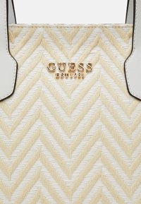 Gros plan d'un motif chevron beige et blanc tissé sur un sac Guess avec un logo doré et des poignées en cuir blanc.