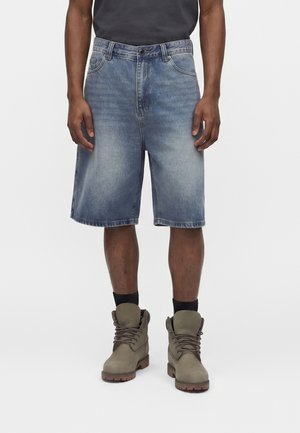 Mężczyzna ubrany w wyblakłe niebieskie krótkie spodnie dżinsowe, czarne skarpetki i jasnobrązowe wiązane buty, stojący na jednolitym jasnym tle.