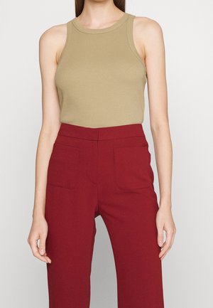 Vrouw draagt mouwloze beige top en rood broek met hoge taille en voorzakken, staand tegen een effen lichte achtergrond.