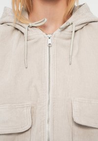 Ελαφρύ γκρι σακάκι από κορδούρα με κουκούλα, μπροστινό φερμουάρ και δύο πλαϊνές τσέπες. Διαθέτει λεία υφή και κορδόνια για ρύθμιση.