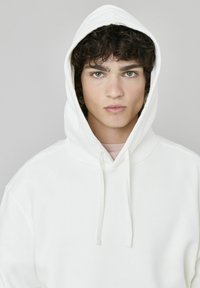 Sweat-shirt à capuche blanc avec une texture douce et des cordons de serrage. Le modèle a des cheveux noirs et bouclés et une expression neutre, avec des yeux bleus.