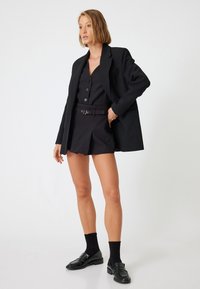 Blazer negro estructurado sobre un chaleco oscuro y pantalones cortos a juego, con botones, un cinturón con una hebilla plateada y mocasines negros con calcetines.