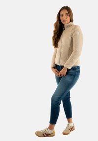 Giacca in pile beige con superficie strutturata, chiusura a zip completa e colletto alto. Abbinata a jeans in denim blu e sneaker beige con dettagli in bordeaux.