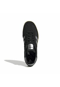 Černý semišový teniska se třemi stříbrnými pruhy, šněrováním, polstrovaným jazykem a texturovanou gumovou podrážkou. Logo je viditelné na jazyku.