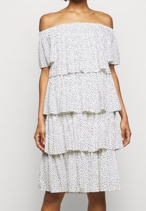 Femme portant une robe blanche à volants et épaules dénudées avec des pois noirs, debout les bras détendus devant un arrière-plan uni.