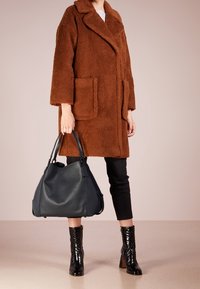 Brązowy, miękki płaszcz z szerokim kołnierzem i naszywanymi kieszeniami, zestawiony z czarnymi spodniami i błyszczącymi botkami. Ciemnoniebieska skórzana torebka.