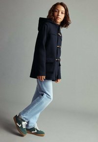 Ung person som bär en mörk huva med knappar, ljusblå jeans och gröna sneakers, som står mot en enkel grå bakgrund.