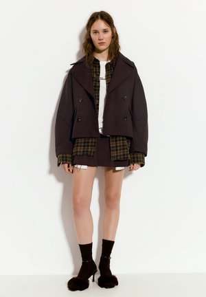 Barna, dupla breasted, rövidített dzseki egy kockás ing felett, rövid szoknyával párosítva. Fekete, magassarkú cipők, szőrmével díszítve, sötét zoknikkal.