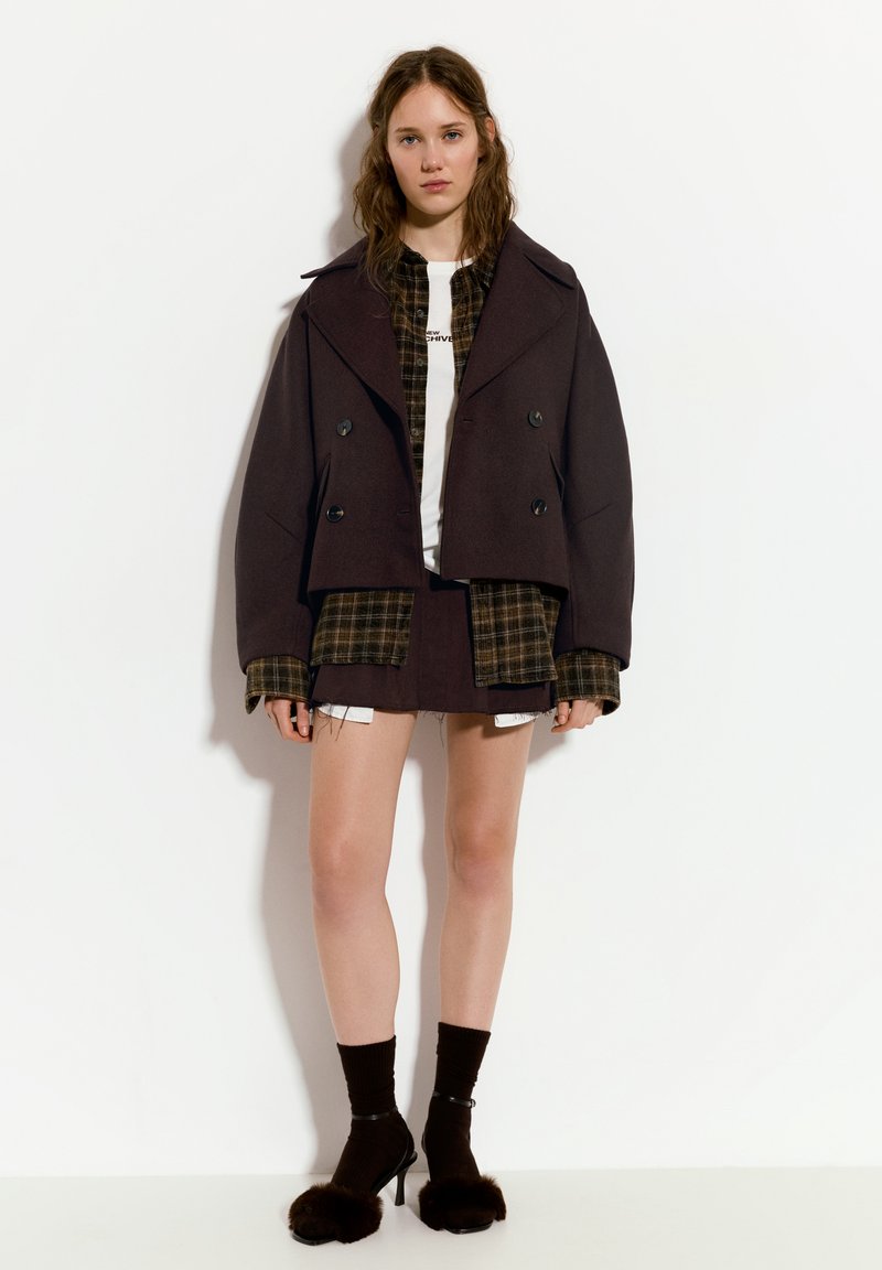 Ruskot beidre sättad, som är över en rutig skjorta, kombinerat med en kort kjol. Svarta högklackade skor med pälsdetaljer och mörka strumpor.